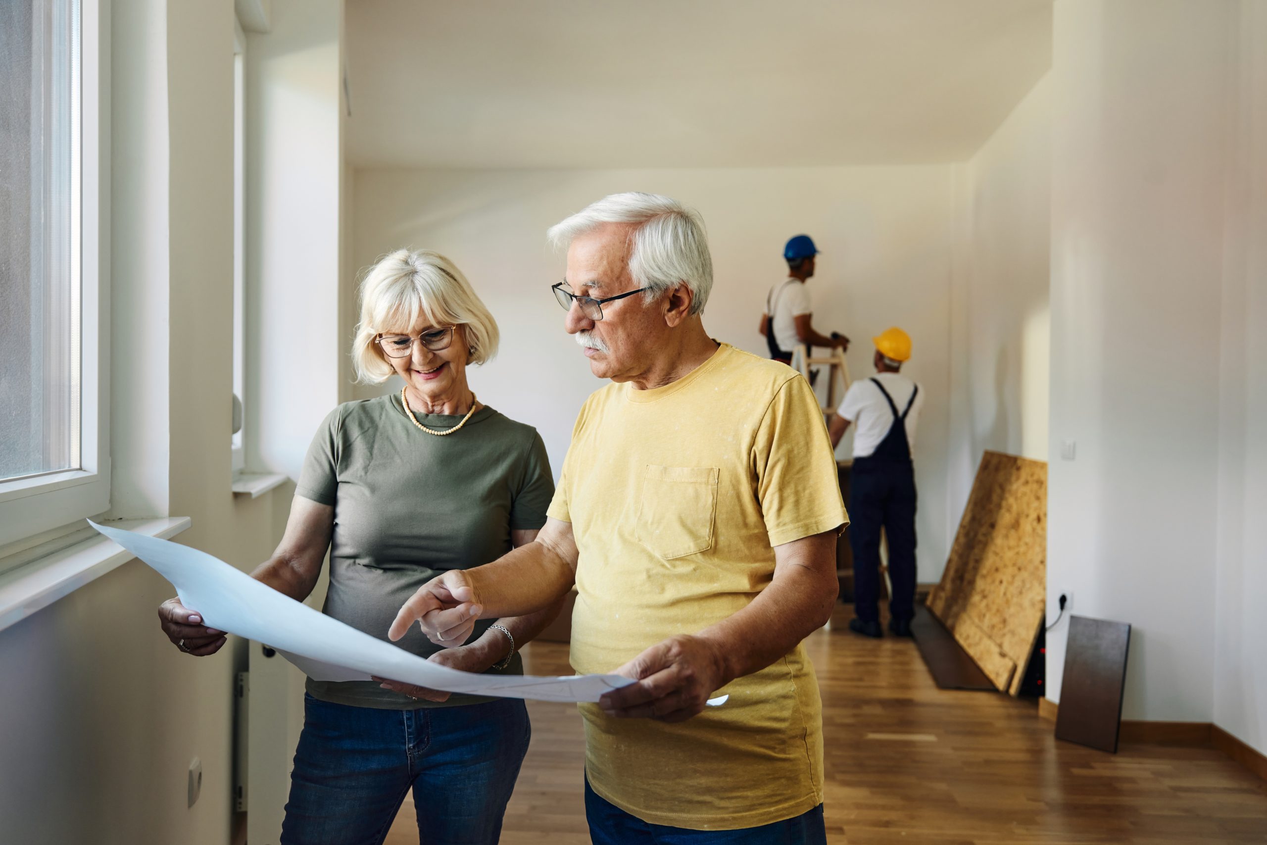 Happy senior couple analyzing housing plans while renovating their apartment. Workers are in the background.