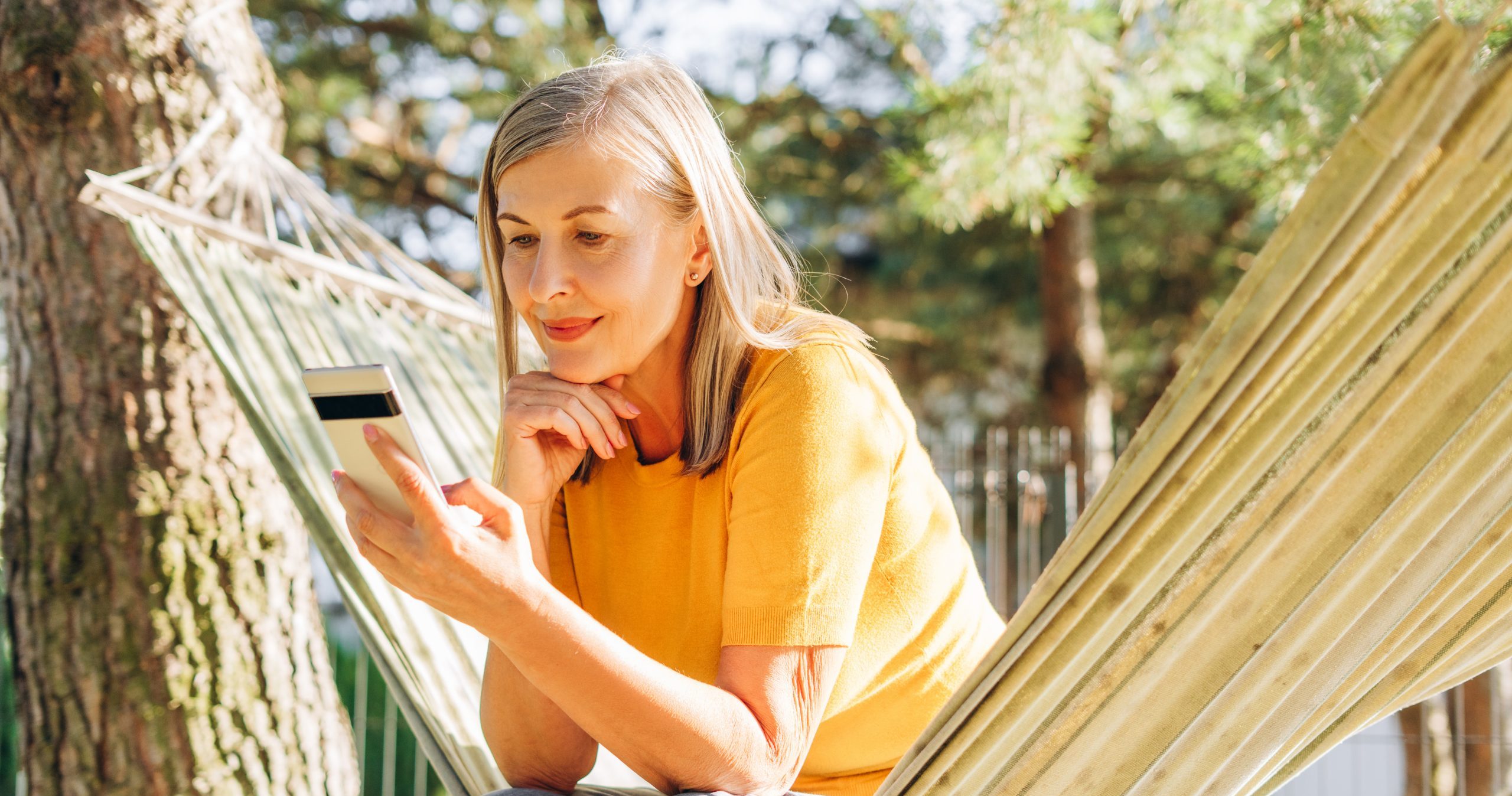Senior adult woman relaxing in a hammock in a garden, happily using her smartphone while enjoying the summer sunshine and nature