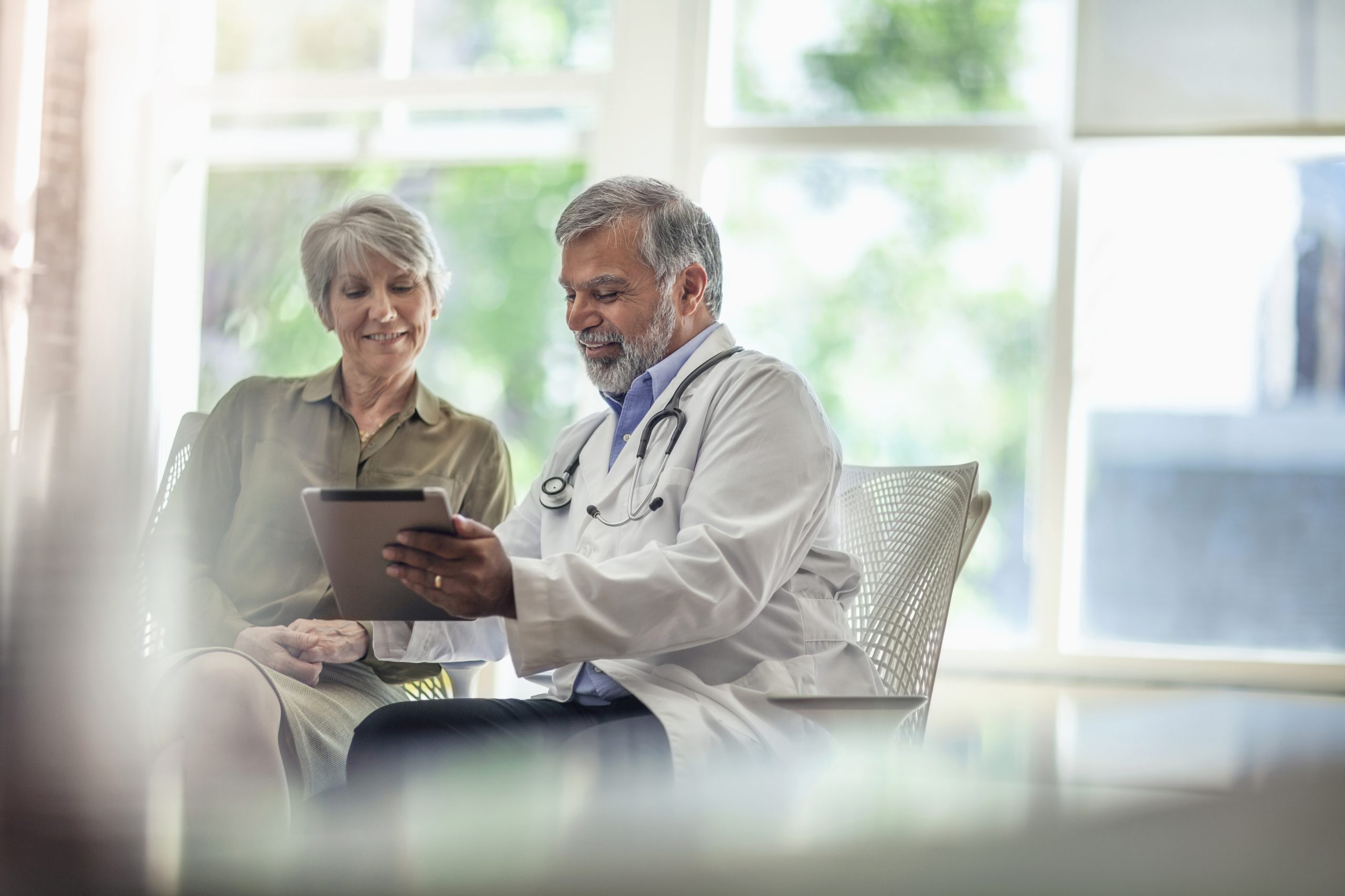 senior man doctor medical professional talks using digital tablet to senior woman in health clinic