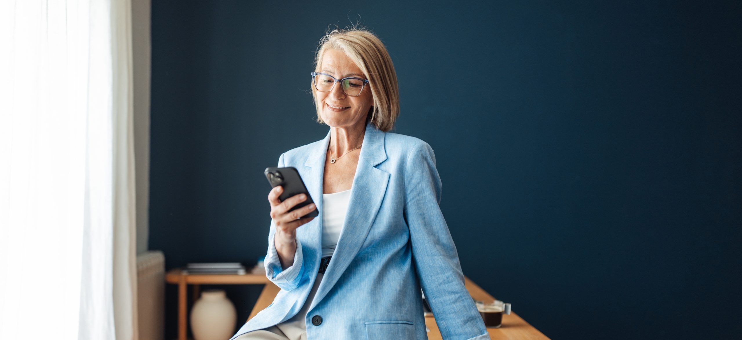 A happy woman leans against a wooden table checking her phone, radiating self-assurance and approachability in a stylish modern workspace filled with natural light.